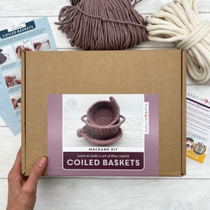 May include: A brown cardboard box with a label that says "Macrame Kit Learn to make a set of three stylish Coiled Baskets" and a picture of three pink coiled baskets. The box is on a white wooden surface.