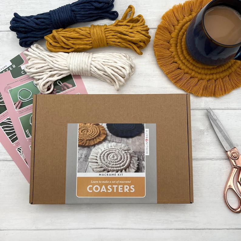 Puede incluir: Una caja de cart&oacute;n marr&oacute;n con una etiqueta que dice "Macrame Kit Learn to make a set of macrame COASTERS" y una imagen de tres posavasos de macram&eacute;. La caja est&aacute; sobre una superficie de madera blanca con tres ovillos de hilo, un par de tijeras y un posavasos de macram&eacute; terminado.