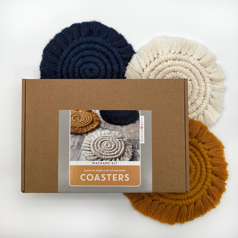 Puede incluir: Un kit de posavasos de macram&eacute; con tres posavasos terminados en azul marino, crema y amarillo mostaza. La caja presenta el texto "Macrame Kit" y "Coasters".