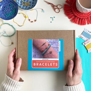 May include: A brown box with a blue and white label that says "Beading Learn to make four colourful stacking Bracelets" with a picture of a person's wrist wearing four bracelets with turquoise and orange beads.