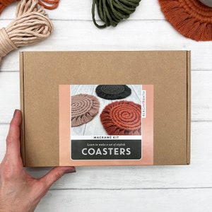 Puede incluir: Una caja de cartón marrón con una etiqueta que dice "Macrame Kit Aprende a hacer un juego de elegantes posavasos COASTERS" y una imagen de tres posavasos de macramé en marrón, beige y negro.