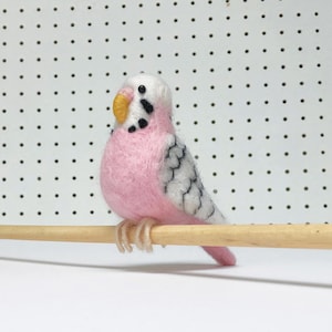 May include: A pink and white needle felted budgie perched on a wooden dowel. The budgie has black eyes and a yellow beak.