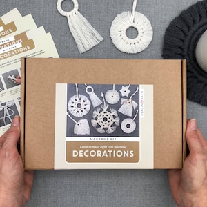 May include: A brown cardboard box with a white label that says "Macrame Kit Learn to make eight cute macrame Decorations" and the logo "Bergin Bath". The box contains eight white macrame ornaments, including a snowflake, a star, and a circle.