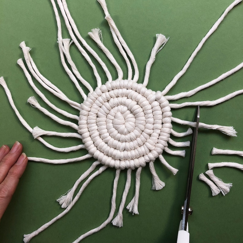 Puede incluir: Un posavasos de macram&eacute; blanco con un dise&ntilde;o circular y bordes con flecos. El posavasos est&aacute; parcialmente terminado y tiene hebras sueltas de hilo.