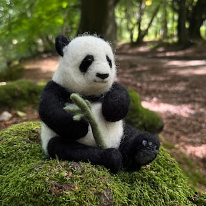 May include: A black and white panda bear plush toy sitting on a bed of green moss. The panda is holding a green bamboo shoot in its paws.