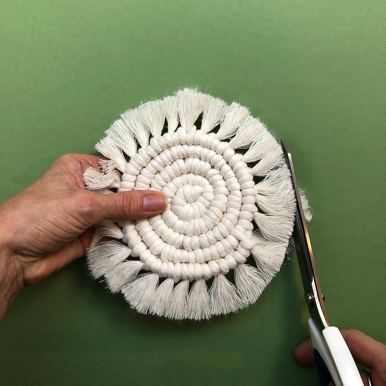 Puede incluir: Un posavasos blanco tejido a crochet con un borde de flecos se est&aacute; recortando con unas tijeras.