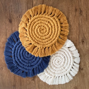 Puede incluir: Tres posavasos redondos de macramé con flecos. Los posavasos son en tonos amarillo, azul y blanco.