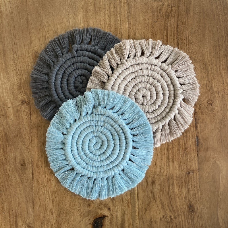 Puede incluir: Tres posavasos de macram&eacute; hechos a mano en azul, gris y beige. Cada posavasos presenta un dise&ntilde;o circular con un borde de flecos.