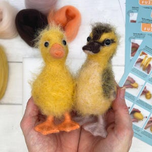 May include: Two handcrafted yellow ducklings with orange beaks and feet are held in a person's hands. One duckling is solid yellow, while the other has brown and yellow markings. Craft supplies, including wool and instructions, are visible in the background.