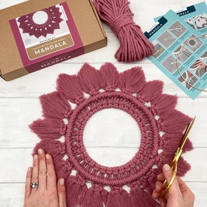 May include: A berry pink macrame mandala kit, including a completed mandala, cord, and instructions. The mandala features a circular design with a fringed edge. Gold scissors are also visible.