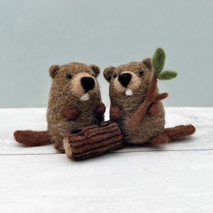 May include: Two brown felted beaver figurines with black noses and white teeth. One beaver holds a small branch with green leaves. A small log is between the beavers. The figurines are on a white surface.