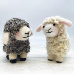 May include: Two handcrafted felt sheep ornaments. One is grey, the other is cream, both with smiling faces and black eyes. The sheep have pink accents on their ears and cheeks. Ideal for a nursery or farm-themed decoration.