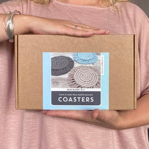 May include: A brown cardboard box with a label that says "Macrame Kit Learn to make these stylish macrame Coasters" and shows three macrame coasters in grey, blue, and pink.