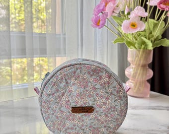 May include: A round, floral-patterned pouch in shades of light blue, pink, and white. The pouch features a zipper closure and a small brown tag with text. The background includes a vase of pink flowers and a window.