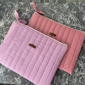May include: Two quilted fabric pouches in pink and red gingham patterns. Each pouch has a white zip and a small brown label. The pink pouch has a matching fabric pull, while the red pouch has a fabric pull with a leaf-like design.