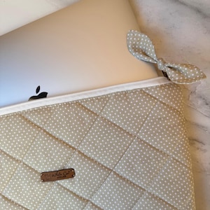 May include: Beige laptop sleeve with white polka dots and a quilted design. The sleeve features a white zip and a bow-shaped pull tab. A laptop is partially visible. The label reads "By White Cat Atelier".