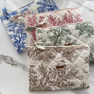 K&ouml;nnte beinhalten: Eine Sammlung gesteppter Kosmetiktaschen in verschiedenen Farben, darunter Blau, Rot, Gr&uuml;n und Braun. Jede Tasche hat ein anderes Toile-Muster und eine passende Stoffschleife am Rei&szlig;verschluss. Die Taschen sind auf einer wei&szlig;en Marmoroberfl&auml;che angeordnet.