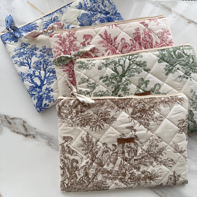K&ouml;nnte beinhalten: Eine Sammlung gesteppter Kosmetiktaschen in verschiedenen Farben, darunter Blau, Rot, Gr&uuml;n und Braun. Jede Tasche hat ein anderes Toile-Muster und eine passende Stoffschleife am Rei&szlig;verschluss. Die Taschen sind auf einer wei&szlig;en Marmoroberfl&auml;che angeordnet.
