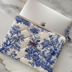May include: Quilted laptop sleeve featuring a blue and white toile design. The sleeve has a beige zip and a matching fabric pull. The pattern depicts scenes of trees, figures, and buildings. A small brown leather tag is attached.