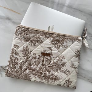 K&ouml;nnte beinhalten: Cremefarbene, gesteppte Laptoptasche mit braunem Toile-de-Jouy-Muster mit Figuren in einer Landschaft. Die Tasche hat einen Rei&szlig;verschluss und eine kleine Stoffschlaufe. Ein silberner Laptop ist teilweise sichtbar.