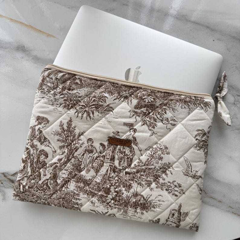 K&ouml;nnte beinhalten: Cremefarbene, gesteppte Laptoptasche mit braunem Toile-de-Jouy-Muster mit Figuren in einer Landschaft. Die Tasche hat einen Rei&szlig;verschluss und eine kleine Stoffschlaufe. Ein silberner Laptop ist teilweise sichtbar.