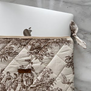 K&ouml;nnte beinhalten: Eine cremefarbene Laptop-H&uuml;lle mit einem braunen Toile-de-Jouy-Muster mit Figuren und Landschaften. Die gesteppte H&uuml;lle hat einen Rei&szlig;verschluss und eine passende Stoffschleife. Ein silbernes Apple-Laptop ist teilweise eingesetzt.