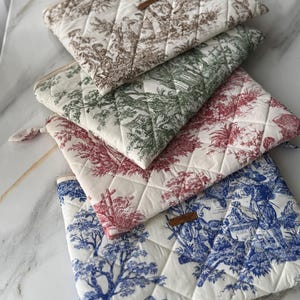 K&ouml;nnte beinhalten: Eine Ansammlung gesteppter Stoffbeutel mit Toile-de-Jouy-Muster. Die Beutel sind in Braun-, Gr&uuml;n-, Rot- und Blaut&ouml;nen auf wei&szlig;em Hintergrund gehalten. Jeder Beutel hat einen Rei&szlig;verschluss und ein kleines Lederetikett.