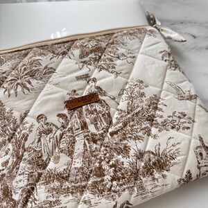 K&ouml;nnte beinhalten: Gesteppte Toilettentasche in Elfenbein und Braun mit Rei&szlig;verschluss und kleiner Stoffschlaufe. Die Tasche hat ein klassisches Toile-Muster, das Figuren in einer Landschaft darstellt. Ein kleines Lederetikett lautet "Fly White Cat Atelier".