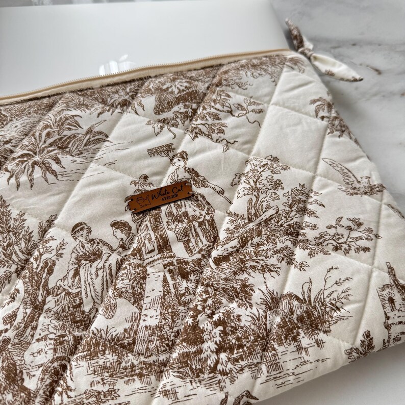 K&ouml;nnte beinhalten: Gesteppte Toilettentasche in Elfenbein und Braun mit Rei&szlig;verschluss und kleiner Stoffschlaufe. Die Tasche hat ein klassisches Toile-Muster, das Figuren in einer Landschaft darstellt. Ein kleines Lederetikett lautet "Fly White Cat Atelier".