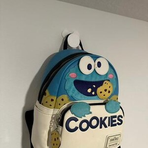 以下が含まれることがあります： クッキーモンスターのデザインが施された青と白のバックパック。バックパックには、ジッパー付きのメインコンパートメント、上部のハンドル、前面ポケットがあり、「COOKIES」の文字がプリントされています。クッキーのチャームがジッパーに付いています。