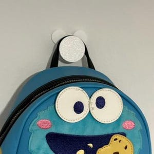 以下が含まれることがあります： クッキーモンスターのデザインのライトブルーのバックパック。大きな白い目とクッキーを持った笑顔の口が特徴です。バックパックは、黒いストラップが付いた白くキラキラ光るフックに吊り下げられています。