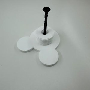 以下が含まれることがあります： ミッキーマウスの頭の形をした白い3Dプリントオブジェクト。白いディスクのスタックと黒いネジが付いています。このオブジェクトはネジを固定するように設計されています。白い表面に置かれています。