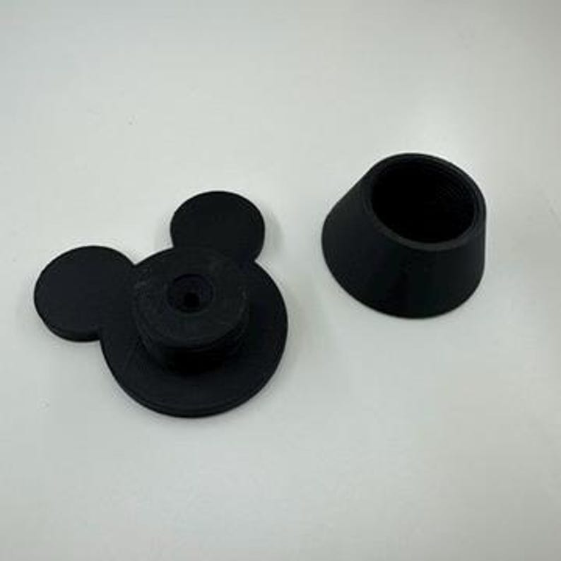以下が含まれることがあります： 黒色の3Dプリントボトルストッパーセット。片方はミッキーマウスの頭の形で、2つの円形の耳と中央の円盤があります。もう片方は、内部にネジ山が切られた円錐形のスリーブです。