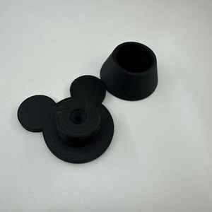 以下が含まれることがあります： 黒い3Dプリントのミッキーマウス型ボトルストッパーと、お揃いの黒い円錐形のトップ。ストッパーは、2つの小さな円形の耳と中央のネジ機構を備えた円形のベースが特徴です。円錐形のトップは、ベースに合うように設計されています。