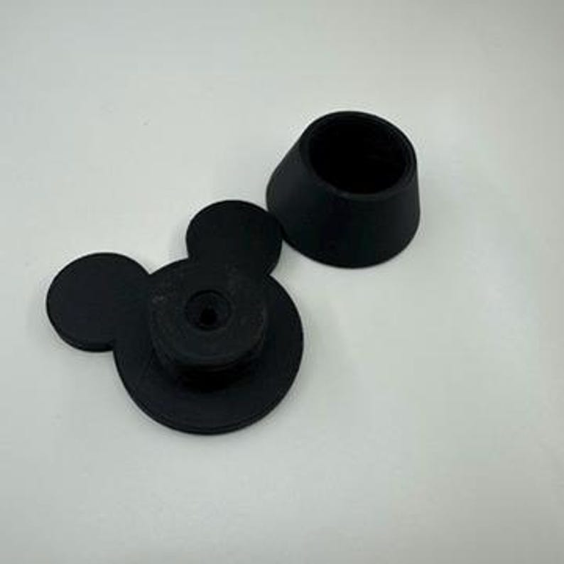 以下が含まれることがあります： 黒い3Dプリントのミッキーマウス型ボトルストッパーと、お揃いの黒い円錐形のトップ。ストッパーは、2つの小さな円形の耳と中央のネジ機構を備えた円形のベースが特徴です。円錐形のトップは、ベースに合うように設計されています。