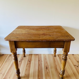 May include: A rectangular wooden table with a light brown finish. The table has a single drawer and four turned legs. The tabletop is made of planks of wood. The table is a vintage or antique piece.