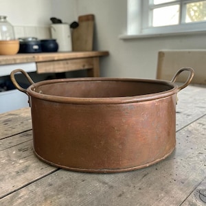 May include: A round copper cooking pot with a flat base and straight sides. The pot features two curved metal handles attached to opposite sides of the rim. The exterior surface shows a weathered, metallic copper finish with visible signs of use.