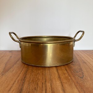 May include: A round, polished brass bowl featuring two curved handles attached to opposite sides. The bowl has a smooth, metallic gold-toned surface and a flat base, presenting a simple, functional design with a consistent finish throughout the exterior.