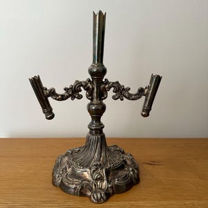 May include: A silver-toned metal candelabra featuring a central pillar and two side arms, each designed to hold a single candle. The base and arms are decorated with intricate embossed scrollwork and floral patterns, with a polished finish.