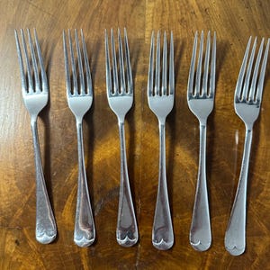 May include: A set of six silver-toned metal forks arranged in a row. Each fork features four long, slender tines and a smooth, tapered handle that ends in a distinct, rounded, scalloped decorative detail at the base of the handle.