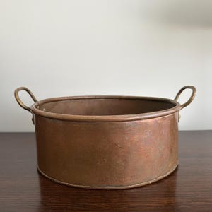 May include: A copper-coloured, oval-shaped metal bowl with two small, curved handles. The bowl has a slightly aged appearance, with visible wear and a rich, warm tone. This vintage-style bowl could be used for decorative purposes or as a small planter.