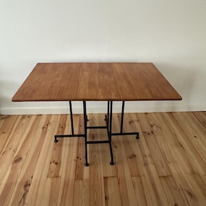 May include: A rectangular wooden table with a light brown finish. The table has a folding design and is supported by black metal legs. The table is approximately 122 cm long and 76 cm wide.