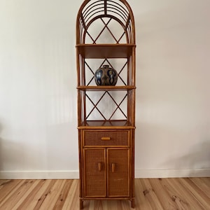 May include: A tall, brown rattan etagere with an arched top and multiple shelves. The etagere features a cabinet with two doors and a drawer. A decorative vase with a blue and brown pattern sits on one of the shelves.