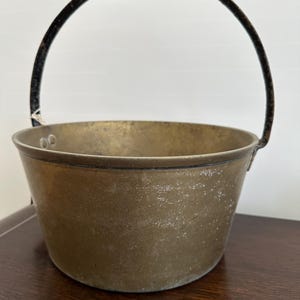 May include: A vintage brass pot with a black metal handle. The pot has a worn, aged look and is perfect for a rustic kitchen or farmhouse decor.