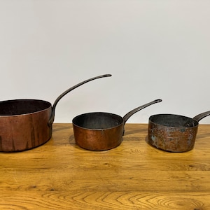 May include: Three antique copper saucepans with long, curved handles. The pans vary in size, with a dark, aged patina. The copper has a rich, reddish-brown hue, and the interiors show signs of use. The pans are arranged on a wooden surface.