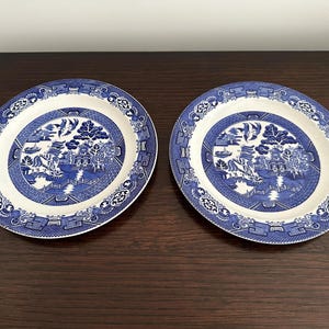 May include: Two blue and white Willow pattern plates. Each plate has a central landscape scene with a bridge, trees, and birds. The rims have a decorative border. The plates are on a dark brown surface.
