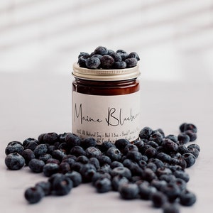 May include: A brown glass candle jar with a gold lid. The jar is filled with a brown candle and has a white label that reads "Maine Blueberry". The label also includes the text "100% All Natural Soy - Net 1.75oz - Hand Poured in Maine". Blueberries are scattered around the jar.