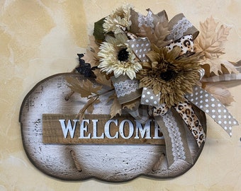 Könnte beinhalten: Ein rustikales, weiß getünchtes Schild in Form eines Kürbisses mit dem Wort "WELCOME" in weißen Buchstaben. Das Schild ist mit einer braunen, weißen und leopardenfarbenen Schleife mit Herbstblättern verziert.