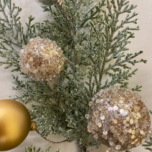 May include: A close-up of a green artificial Christmas tree branch with three ornaments. Two of the ornaments are gold and covered in glitter. The third ornament is silver and shiny.