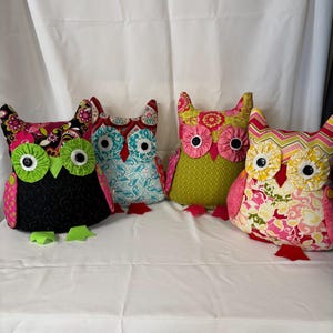 May include: Four colorful owl-shaped pillows. Each owl has large button eyes and a unique patterned fabric body. The owls are in shades of pink, green, black, and blue, with various floral and geometric designs. The pillows are arranged on a white surface.
